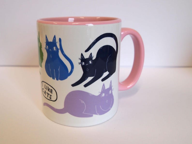 May include: A white ceramic mug with a pink rim and handle. The mug features a colorful illustration of cats with the text "I like cats" in a speech bubble.