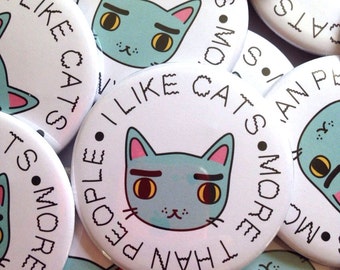 Op de afbeelding: Een verzameling ronde buttons met de illustratie van een lichtblauwe kat met gele ogen en roze oren. De tekst "I LIKE CATS MORE THAN PEOPLE" is in een zwart, handgetekend lettertype rondom het gezicht van de kat gedrukt.