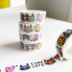 Peut inclure: Cinq rouleaux de ruban adh&eacute;sif washi empil&eacute;s avec des motifs de chats et de c&oelig;urs color&eacute;s. Le ruban est d&eacute;roul&eacute; et affiche le texte "J'aime les chats".
