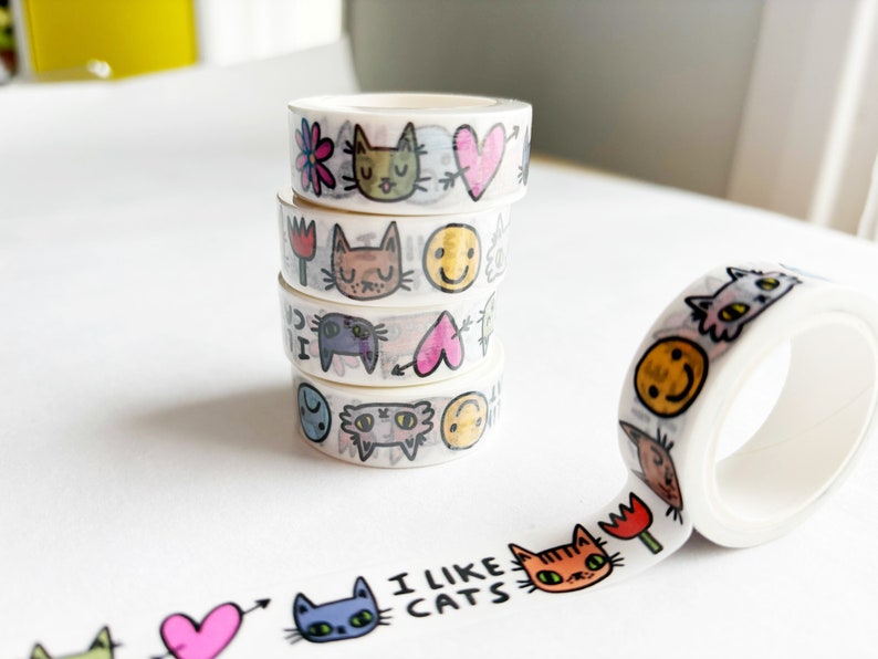 Peut inclure: Cinq rouleaux de ruban adh&eacute;sif washi empil&eacute;s avec des motifs de chats et de c&oelig;urs color&eacute;s. Le ruban est d&eacute;roul&eacute; et affiche le texte "J'aime les chats".