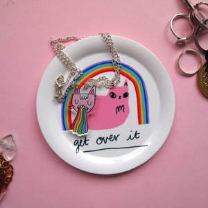 May include: A white ceramic dish with a rainbow and two pink cats with the text "Get over it". The dish has a silver chain necklace with a pink cat charm.