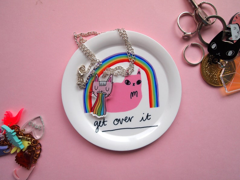May include: A white ceramic dish with a rainbow and two pink cats with the text "Get over it". The dish has a silver chain necklace with a pink cat charm.