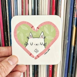 Pode incluir: Um descanso de copo de cerâmica branca com um design em forma de coração rosa que apresenta dois vermes de desenho animado e um gato branco com o texto "Eu gosto de gatos" em letras pretas.