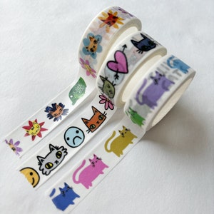 Peut inclure: Cinq rouleaux de ruban adh&eacute;sif washi color&eacute; avec des motifs de chats et de fleurs. Le ruban est blanc avec des chats et des fleurs de diff&eacute;rentes couleurs. Le texte sur l'un des rubans dit "J'aime le g&acirc;teau".