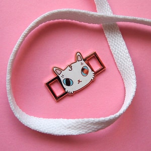 Op de afbeelding: Een witte kattenkopbedel met blauwe ogen en een roze neus, bevestigd aan een gouden metalen schuif. De schuif zit op een wit veterschoen.