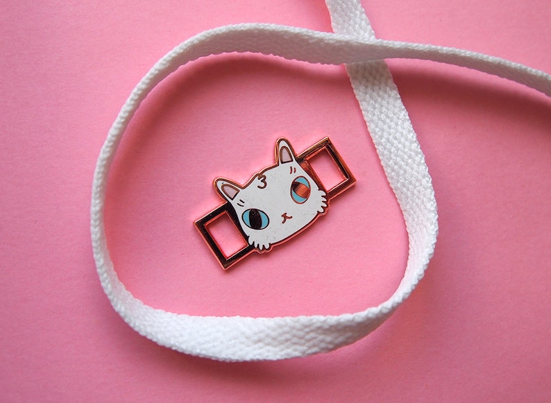 Op de afbeelding: Een witte kattenkopbedel met blauwe ogen en een roze neus, bevestigd aan een gouden metalen schuif. De schuif zit op een wit veterschoen.