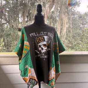 May include: A dark gray t-shirt featuring a graphic of a skull wearing a baseball cap with the text FALL OUT BOY and FOB, along with the phrase WE ARE THE POISONED. The shirt has green and orange quilted fabric panels attached to the sides.
