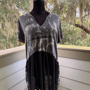 May include: A grey and black tie-dye t-shirt with a V-neckline featuring the Fender logo and a graphic of an electric guitar with the text SINCE 1946. The lower portion of the shirt has a solid black sheer panel and black and white patterned side panels.