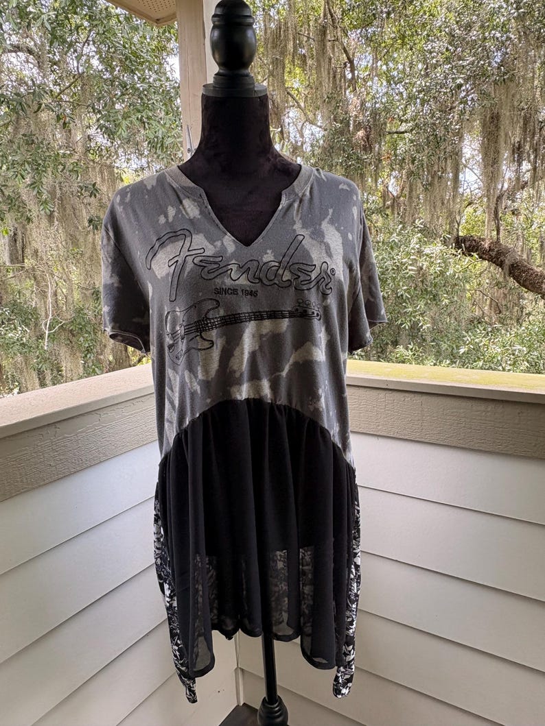 May include: A grey and black tie-dye t-shirt with a V-neckline featuring the Fender logo and a graphic of an electric guitar with the text SINCE 1946. The lower portion of the shirt has a solid black sheer panel and black and white patterned side panels.
