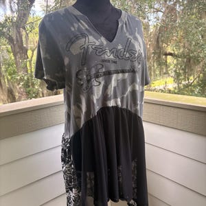 May include: A grey and black tie-dye t-shirt with a V-neckline featuring the Fender logo and a graphic of an electric bass guitar with the text SINCE 1946. The lower portion of the shirt has a solid black sheer panel and a black and white floral print.
