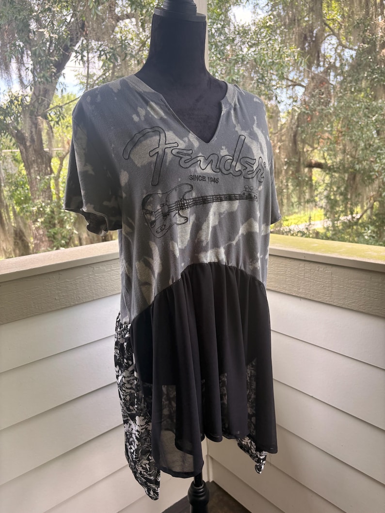 May include: A grey and black tie-dye t-shirt with a V-neckline featuring the Fender logo and a graphic of an electric bass guitar with the text SINCE 1946. The lower portion of the shirt has a solid black sheer panel and a black and white floral print.