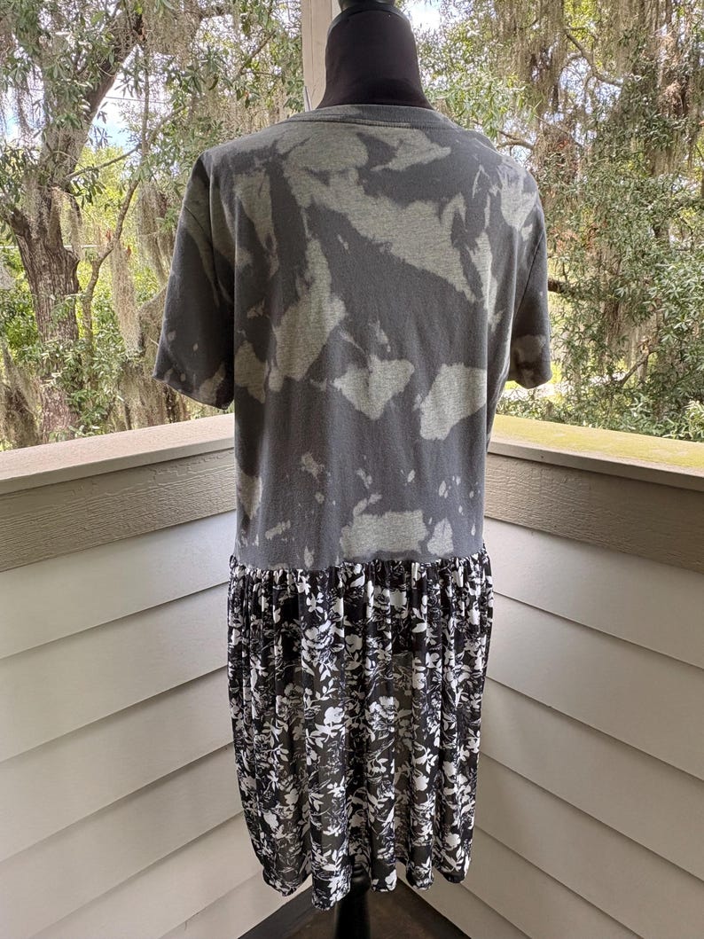 May include: A short-sleeved shirt featuring a charcoal grey and light grey acid-wash pattern on the upper portion, with a contrasting black and white floral print fabric attached at the waist, creating a layered, dress-like appearance.