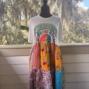 May include: A white t-shirt dress featuring a Grateful Dead graphic with dancing bears and a lightning bolt logo. The skirt portion is constructed from a patchwork of multi-colored fabric panels in shades of yellow, red, blue, and white with various patterns.
