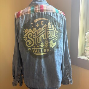 May include: A blue denim shirt with a green and yellow patch on the back that says "Stardew Valley". The patch has a stylized image of a farm with a chicken, mountains, and a crescent moon.