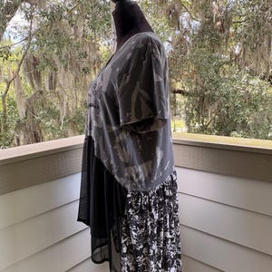 May include: A short-sleeve shirt featuring a charcoal grey bleached tie-dye pattern on the upper section, a solid black sheer fabric panel in the middle, and a black and white floral print fabric panel along the lower back hemline.