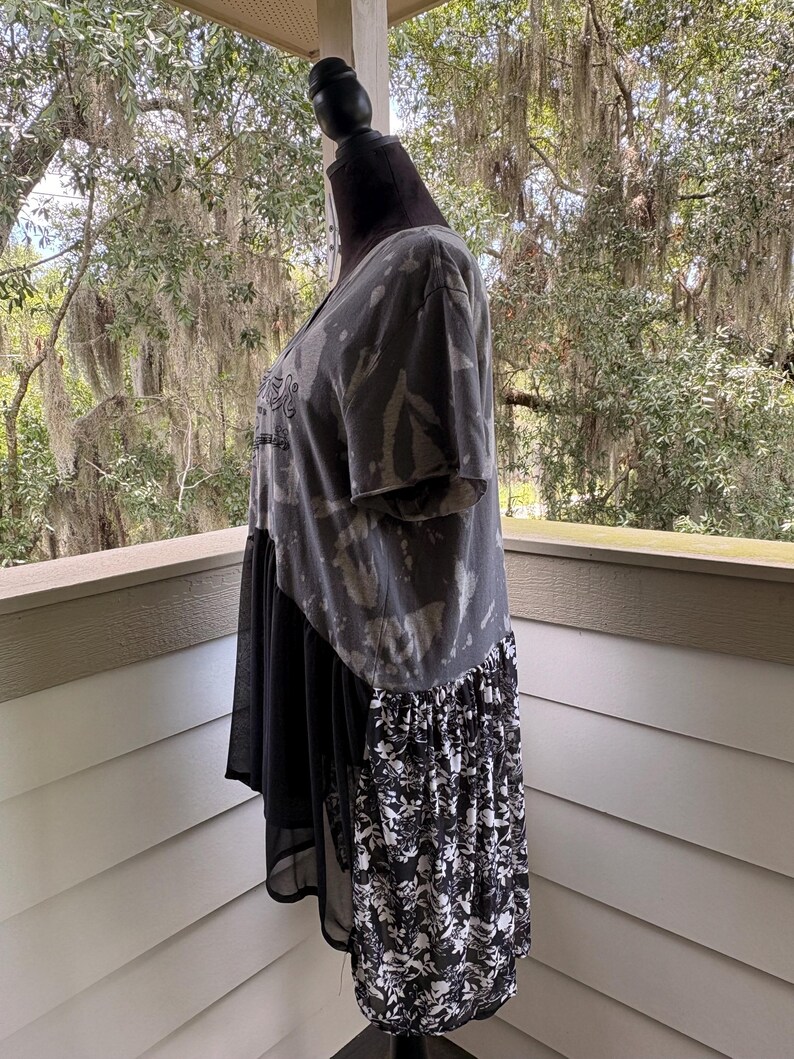 May include: A short-sleeve shirt featuring a charcoal grey bleached tie-dye pattern on the upper section, a solid black sheer fabric panel in the middle, and a black and white floral print fabric panel along the lower back hemline.