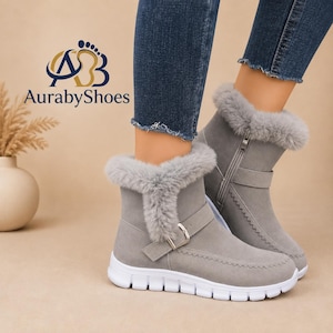 May include: Grey ankle boots with a faux fur collar, a side zipper, and a decorative strap with a silver buckle. The boots feature a white rubber sole and visible stitching along the front. The AurabyShoes logo is visible in the upper left corner.