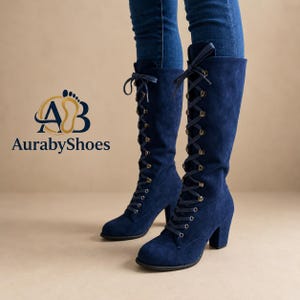 May include: A pair of knee-high navy blue suede boots featuring a lace-up front with dark metal eyelets and matching navy laces tied at the top. The boots have a block heel and are displayed with blue jeans. The logo AurabyShoes is visible.
