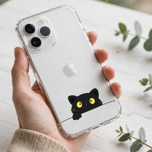 May include: A clear smartphone case featuring a graphic of a black cat with large yellow eyes peeking over a horizontal line. The case is transparent, revealing the silver back of the phone and the Apple logo positioned above the cat illustration.
