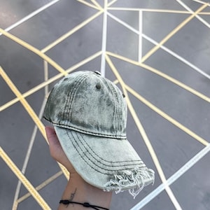 Puede incluir: Una gorra de béisbol color verde salvia con un acabado desgastado y lavado al ácido. La gorra presenta una visera curva con bordes deshilachados, una construcción de seis paneles con ojales cosidos y un botón a juego en la parte superior.