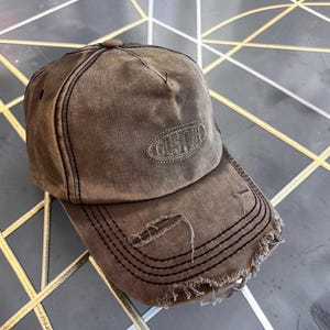 Op de afbeelding: Een bruine baseballpet met een verweerde, versleten afwerking en gerafelde randen aan de gebogen klep. Op het voorpaneel bevindt zich een ovaal embleem met het woord CUSTOM geborduurd in bijpassend bruin garen met zichtbare stiksels.
