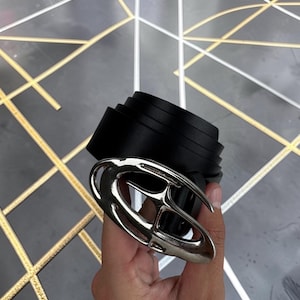 Op de afbeelding: Een zwarte riem met een grote, gepolijste zilverkleurige metalen gesp met een abstract, ovaalvormig uitgesneden ontwerp. De riemband is gemaakt van een glad, matzwart materiaal en is in een cirkelvorm opgerold.