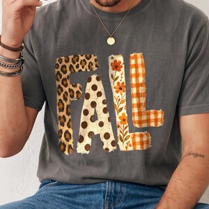 Könnte beinhalten: Ein anthrazitfarbenes Kurzarm-T-Shirt mit dem Wort FALL in großen Buchstaben. Jeder Buchstabe hat ein eigenes Muster: das F ist mit Leopardenmuster, das A mit braunen Punkten, das L mit orangefarbenen Blumen und das L mit orangefarbenem Karomuster.