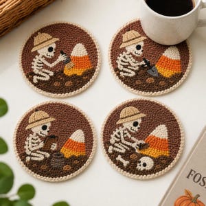 May include: Four round punch needle coasters featuring skeletons wearing pith helmets excavating candy corn. Each coaster has a brown background with white, orange, and yellow details. The skeletons are shown using tools like brushes, shovels, and clipboards to dig up the candy corn pieces.