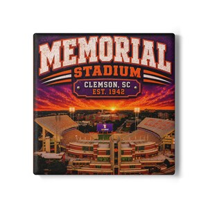 May include: A square wall art print featuring an aerial view of Memorial Stadium in Clemson, SC, established in 1942. The image displays the stadium under a vibrant orange and purple sunset sky with bold white and orange text overlaying the scene.