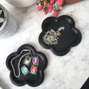 May include: Two black flower-shaped trays made of a matte material. One tray holds a silver-toned chain bracelet with four oval charms featuring colorful religious figures. The other tray contains a silver-toned chain with a small charm.