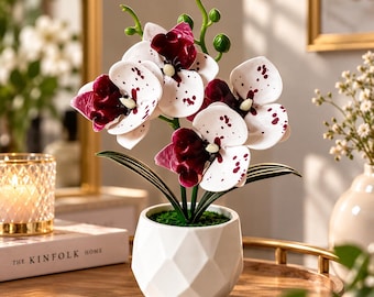 May include: A potted artificial orchid plant featuring white petals with deep burgundy spots and a solid dark burgundy center. The plant is housed in a white, geometric, faceted ceramic pot filled with green moss at the base.