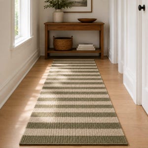 Peut inclure: Un long tapis de couloir rectangulaire présentant un motif à rayures horizontales répétitives dans des tons alternés de vert sauge et de crème. Le tapis a une texture tissée et est placé au centre sur un plancher en bois clair.