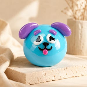 Könnte beinhalten: Eine kugelförmige Glasfigur in Form eines Hundekopfes mit hellblauer Grundfarbe. Sie hat zwei violette Ohren, zwei violette Augenbrauen, weiß-schwarze Augen, eine schwarze Nase und einen Mund sowie eine kleine herausgestreckte rosa Zunge.