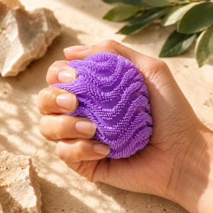 Könnte beinhalten: Ein runder, violetter Silikon-Reinigungsschwamm mit einer dichten Textur aus kleinen, erhabenen und flexiblen Borsten in einem Zickzack-Muster. Das Objekt ist kompakt und so geformt, dass es bequem in die Handfläche passt.