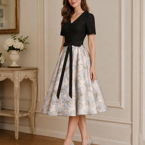 May include: A knee-length dress featuring a black short-sleeved bodice with a V-neckline and a row of buttons. The skirt is a flared, light blue and cream floral jacquard fabric, accented by a solid black ribbon tied at the waist.