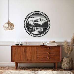 May include: A circular black metal wall art piece featuring a detailed cutout design of a steam locomotive crossing a stone arch bridge, with a backdrop of pine trees and mountain peaks, all enclosed within a decorative geometric lattice border.