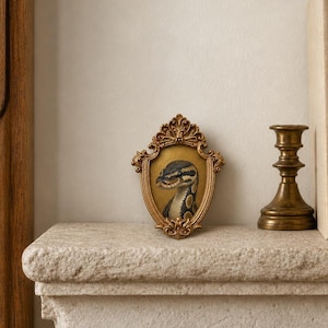 Può includere: Una piccola cornice ovale dorata e ornata contenente un'illustrazione dettagliata di un serpente con squame a motivi marrone chiaro e scuro. La cornice presenta intricati motivi a volute e intagli decorativi sui bordi superiore e inferiore.