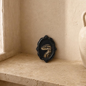 May include: A small, oval-shaped black picture frame featuring an ornate, decorative border. Inside the frame is a detailed illustration of a brown and tan snake with a patterned, scaled body set against a solid black background.