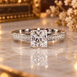 May include: A silver-toned ring featuring a large, square-cut princess diamond at the center. The band is crafted from a polished metal and is encrusted with a row of small, round-cut diamonds along both sides of the central stone setting.