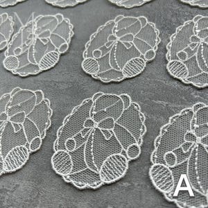 Puede incluir: Múltiples apliques de encaje blanco, cada uno con forma ovalada y un motivo detallado de oso de peluche. El diseño incluye una figura central de oso con pajarita, rodeada por un borde festoneado. La letra A es visible en la esquina inferior derecha.