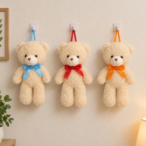 Op de afbeelding: Drie beige pluche teddyberen met krullende vacht, elk hangend aan een witte muurhaak met een lintlus. De beren hebben bruine neuzen en satijnen strikken in felblauw, rood en oranje, hangend tegen een effen muur.