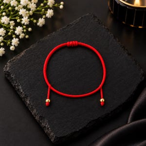 May include: A thin red cord bracelet featuring a sliding knot closure mechanism and two small, polished gold-tone metal beads at the ends of the cord. The bracelet is circular in shape and constructed from a smooth, vibrant red string material.