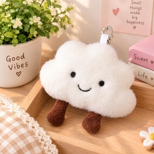 May include: A white plush cloud keychain with a smiling face, black embroidered eyes and mouth, and two brown fuzzy legs. The cloud has a silver metal lobster clasp attached to the top. The texture is soft and fluffy, featuring a rounded shape.