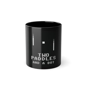 Könnte beinhalten: Ein schwarzer Keramikbecher mit einem minimalistischen Grafikdesign aus zwei vertikalen weißen Balken und einem kleinen weißen Quadrat in der Mitte. Unter der Grafik ist der Text "TWO PADDLES AND A DOT" in einer weißen, verpixelten Blockschrift gedruckt.