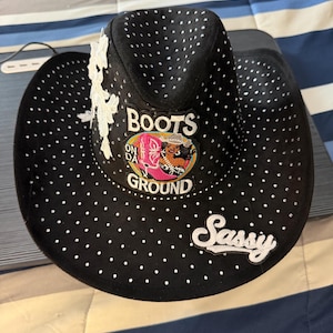 Op de afbeelding: Een zwarte cowboyhoed met witte stippen, een witte kanten bloemapplicatie aan de zijkant en twee patches. Eén patch toont een roze laars met de tekst "BOOTS ON DA GROUND" en de andere patch bevat het woord "Sassy" in wit cursief schrift.