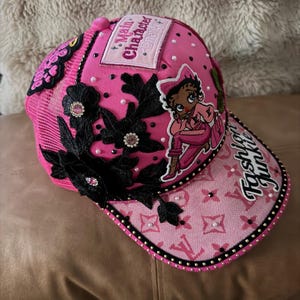May include: A bright pink trucker hat featuring a patch that reads Main Character Energy, a cartoon character graphic, and the text Fashion Junkie on the brim. The hat is decorated with black floral lace appliques, small white pearls, and studs.