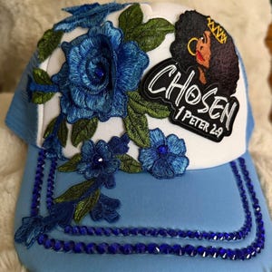 May include: A light blue and white trucker hat featuring embroidered blue roses with green leaves, a patch depicting a person with dark hair wearing a gold crown, and the text "CHOSEN 1 PETER 2:9". The brim is accented with rows of blue rhinestones.