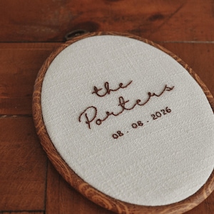 Könnte beinhalten: Ein ovaler Stickrahmen mit cremefarbenem Stoff und dunkelbraun handgesticktem Text, der the Porters und das Datum 08. 08. 2026 zeigt. Der Rahmen hat eine Holzmaserungsoptik und liegt schräg auf einer flachen Oberfläche.