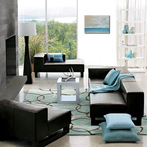 May include: A modern living room featuring a brown leather sectional sofa, a white coffee table, and a blue and white patterned throw. The room has a large window with a view of trees and water. A painting of a seascape hangs on the wall.