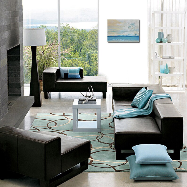 May include: A modern living room featuring a brown leather sectional sofa, a white coffee table, and a blue and white patterned throw. The room has a large window with a view of trees and water. A painting of a seascape hangs on the wall.